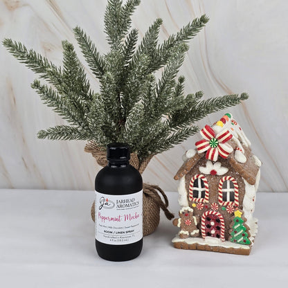 A small artificial pine tree, a Jarhead Aromatics Peppermint Mocha Room/Linen Spray from the Winter Collection, and a decorated gingerbread house sit on a white surface against a marbled background.