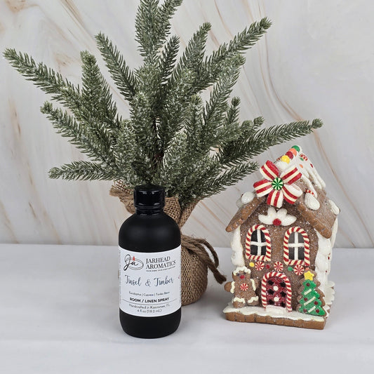 A small artificial pine tree and gingerbread house sit beside a Jarhead Aromatics Tinsel & Timber Room/Linen Spray (Winter Collection), adding a cozy holiday scent to the light, marble-like surface.