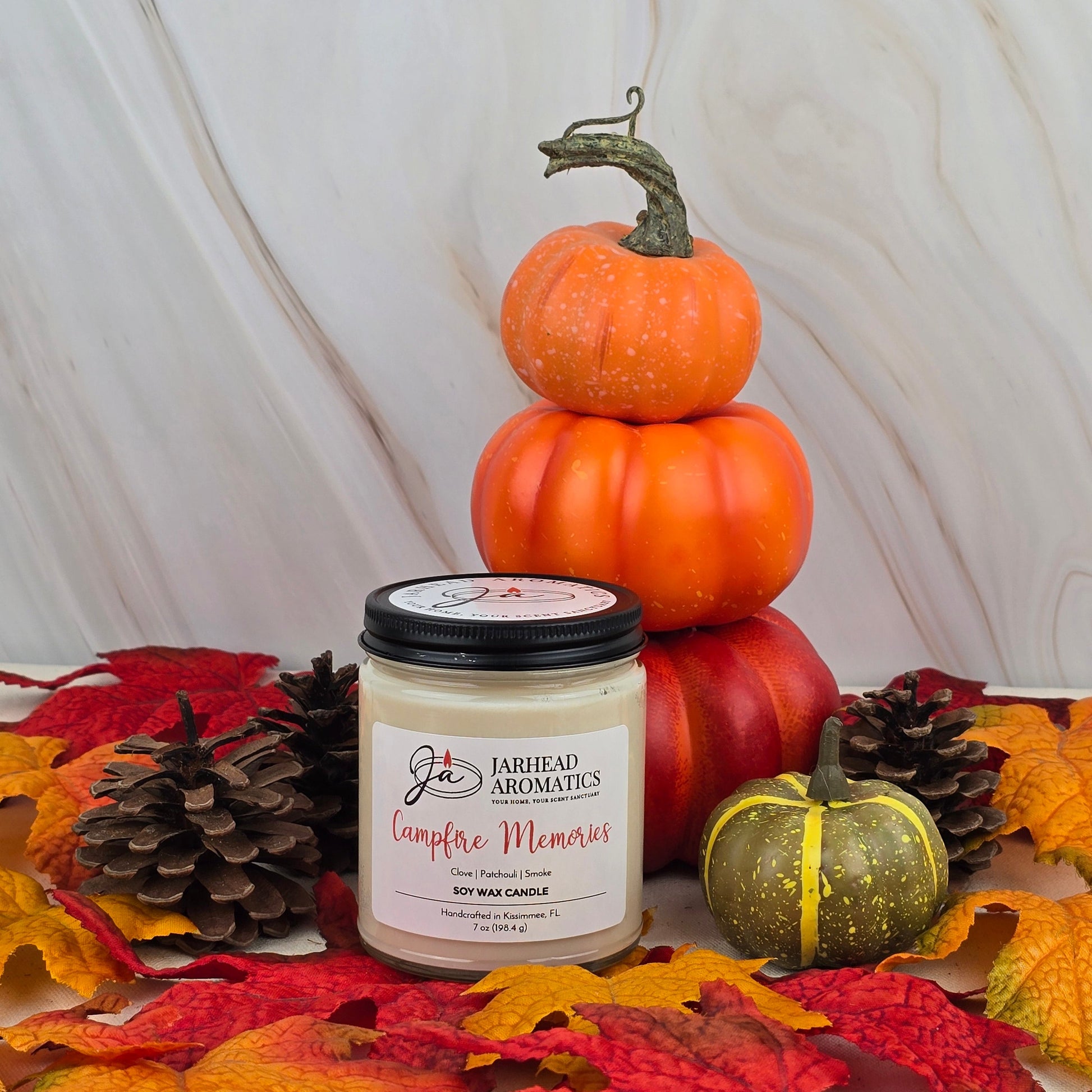 The Campfire Memories Soy Wax Candle from Jarhead Aromatics Fall Collection, with a smoky scent, is displayed among pinecones, autumn leaves, a green pumpkin, and orange pumpkins for a cozy seasonal vibe.