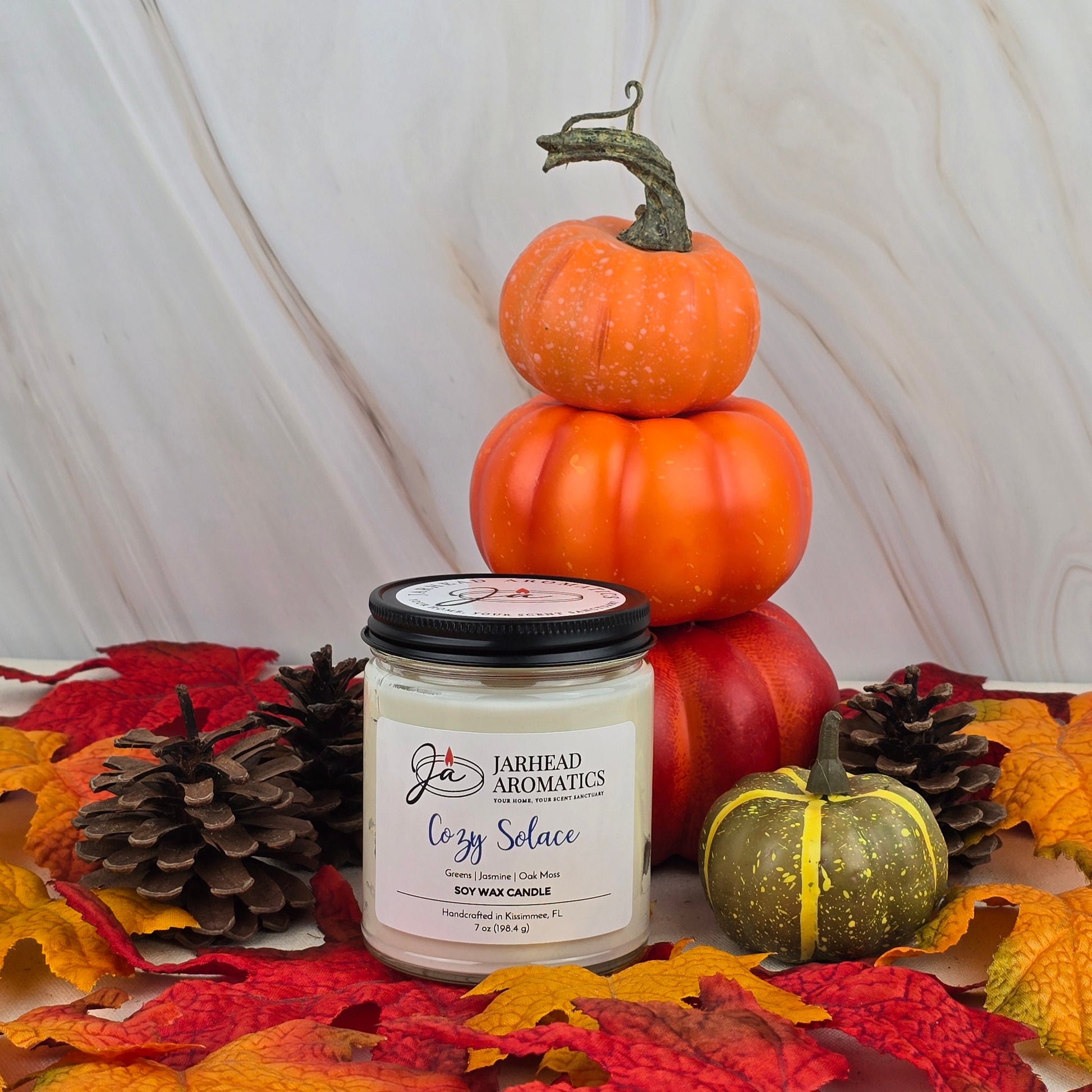The Cozy Solace - Soy Wax Candle from Jarhead Aromatics’ Fall Collection, infused with jasmine, is displayed among autumn leaves, pinecones, a small green pumpkin, and orange pumpkins on a marble background.