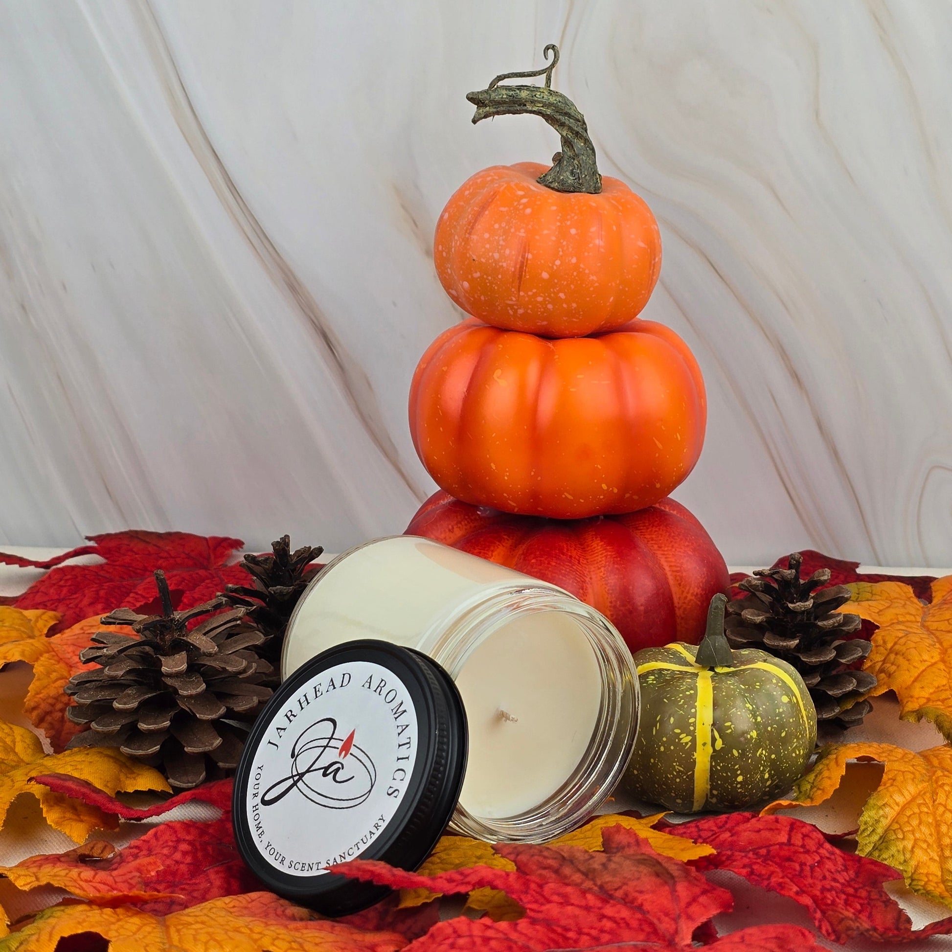 The Cozy Solace soy wax candle from Jarhead Aromatics’ Fall Collection, with its open lid and label, rests among artificial autumn leaves, pinecones, and pumpkins on marble—filling the air with oak moss and fresh greens.