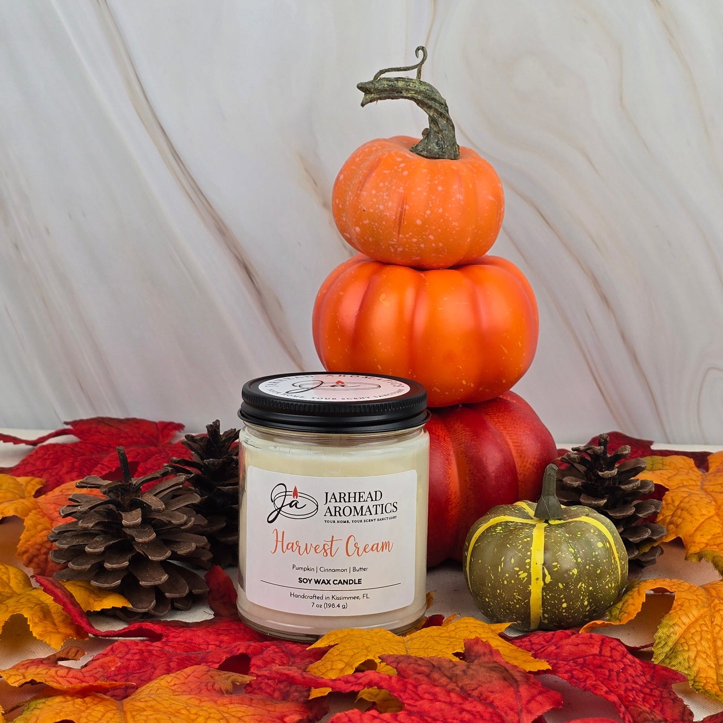 The Jarhead Aromatics Harvest Cream - Soy Wax Candle from the Fall Collection rests on autumn leaves with pinecones, a green-yellow pumpkin, and stacked orange pumpkins, offering a cozy seasonal fragrance against a marble backdrop.