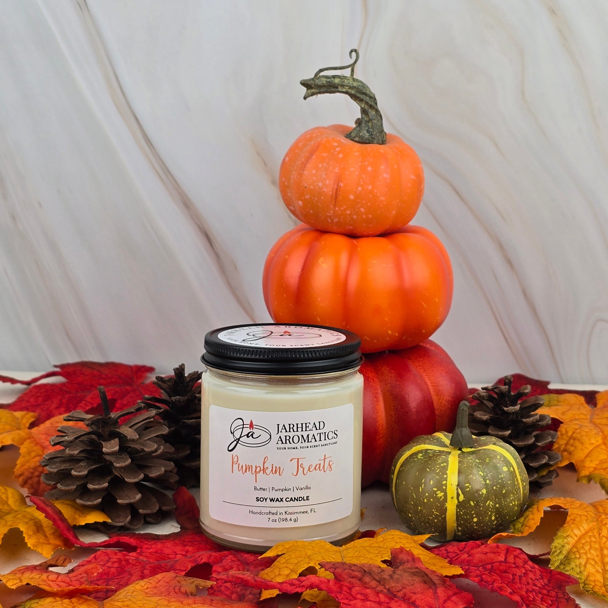 The Pumpkin Treats - Soy Wax Candle from Jarhead Aromatics Fall Collection sits beside a green pumpkin, pinecones, and orange pumpkins, all surrounded by autumn leaves—evoking the perfect fall scent.