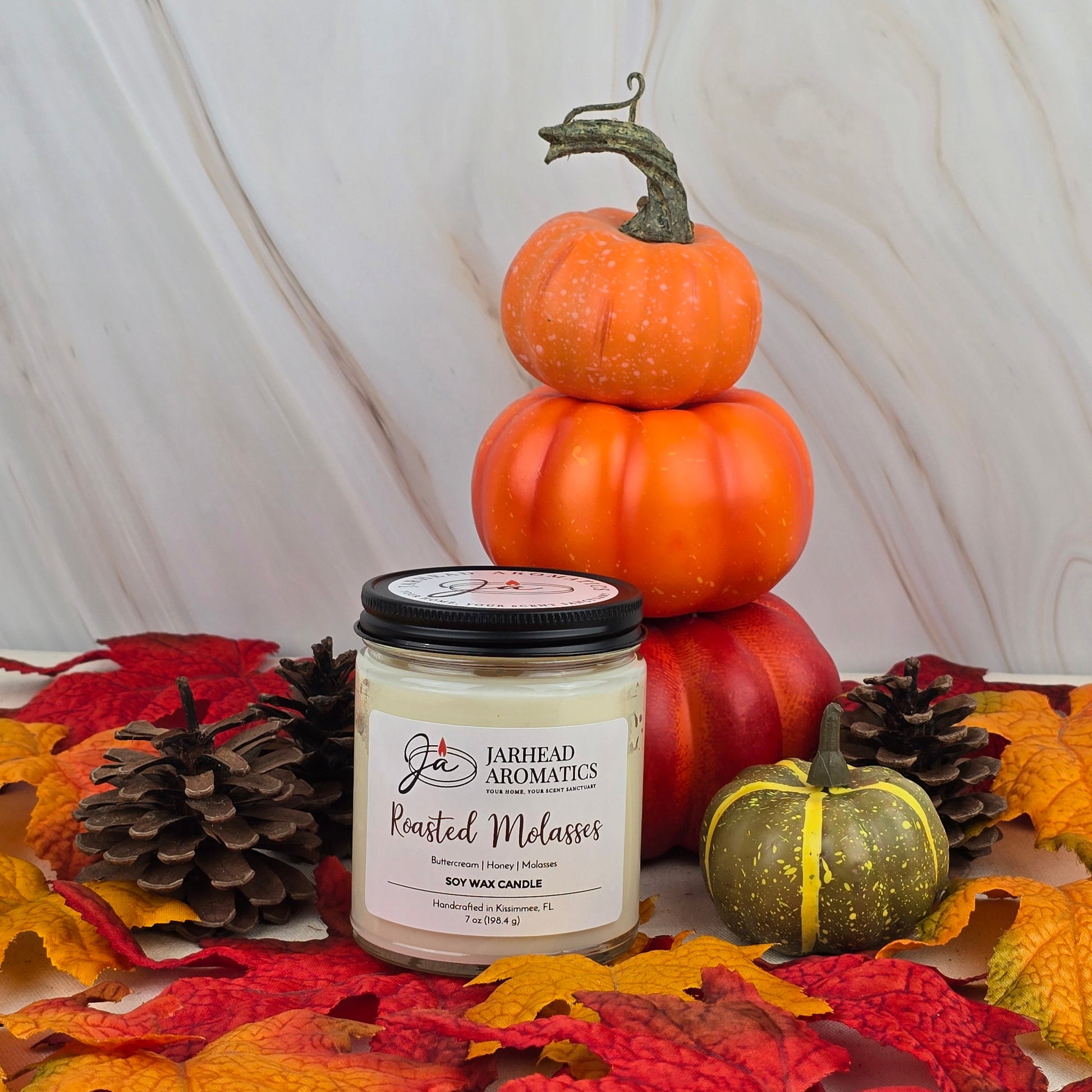A Jarhead Aromatics Roasted Molasses - Soy Wax Candle from the Fall Collection rests on autumn leaves with pinecones, a green pumpkin, and orange pumpkins, infusing the air with cozy caramelized molasses atop a light marble surface.