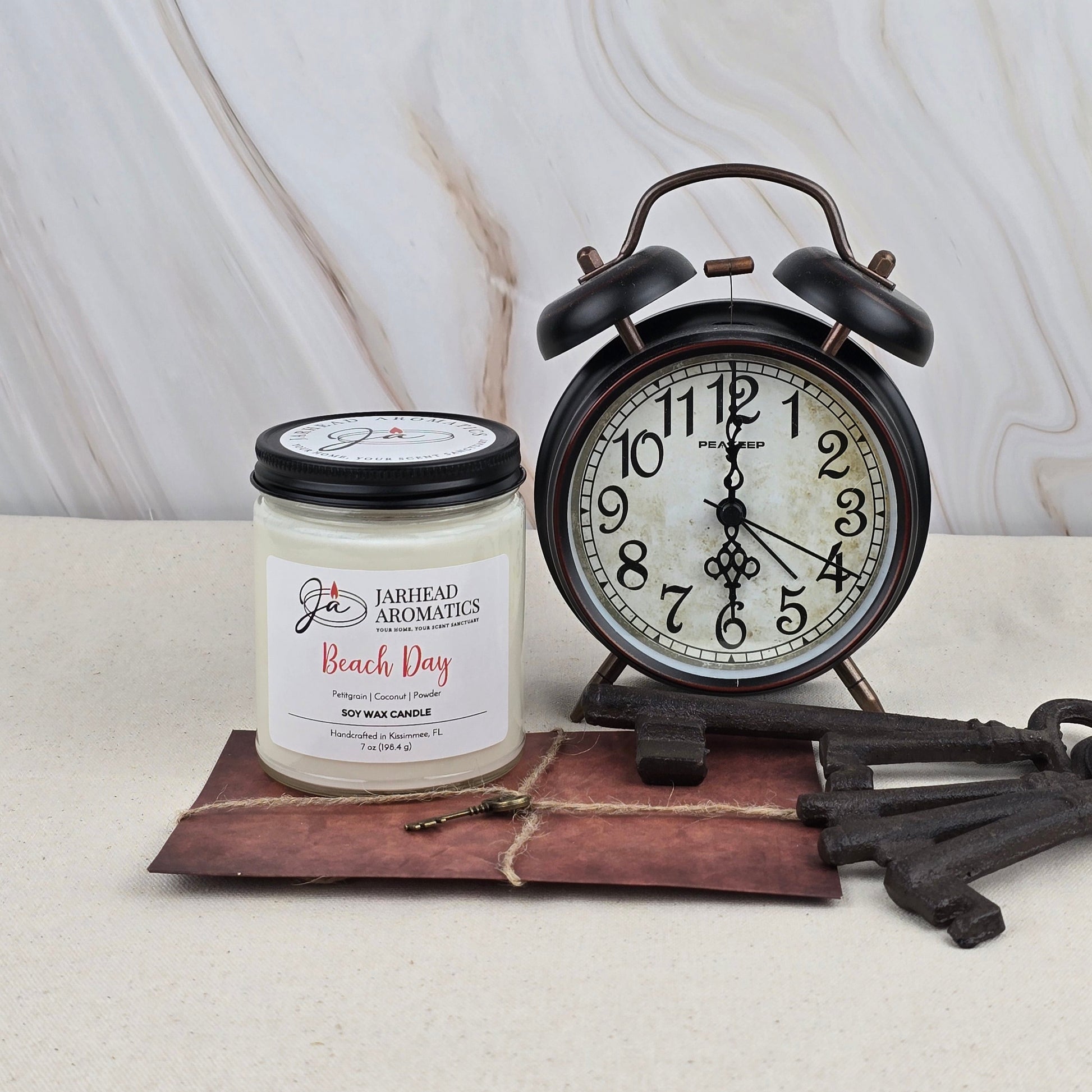 A Jarhead Aromatics Beach Day - Soy Wax Candle with a refreshing beach scent sits beside a black vintage alarm clock and three large old keys on a brown leather mat against a marble-patterned background.
