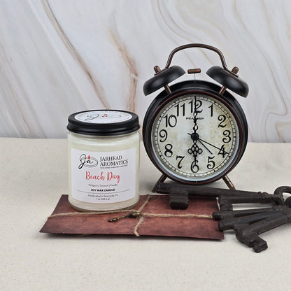 A Jarhead Aromatics Beach Day - Soy Wax Candle with a refreshing beach scent sits beside a black vintage alarm clock and three large old keys on a brown leather mat against a marble-patterned background.
