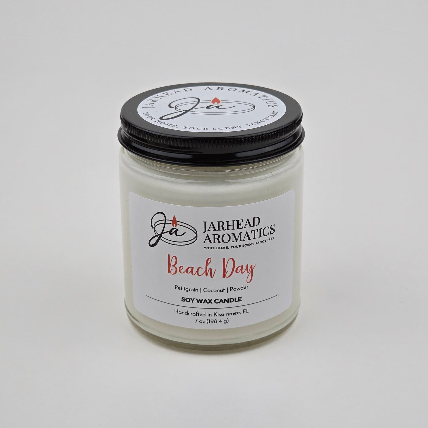 A white jar candle labeled Jarhead Aromatics Beach Day Soy Wax Candle | Coconut, Citrus & Coastal Breeze Candle features a black lid and is handcrafted in Navarre, FL. This candle offers a coastal fragrance with notes of coconut, citrus, and coastal breeze, providing a long-lasting scent perfect for evoking seaside relaxation.