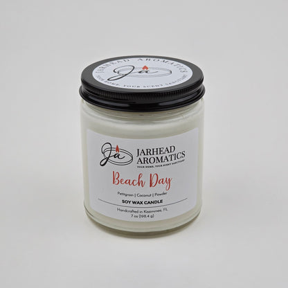 A white jar candle labeled Jarhead Aromatics Beach Day Soy Wax Candle | Coconut, Citrus & Coastal Breeze Candle features a black lid and is handcrafted in Navarre, FL. This candle offers a coastal fragrance with notes of coconut, citrus, and coastal breeze, providing a long-lasting scent perfect for evoking seaside relaxation.