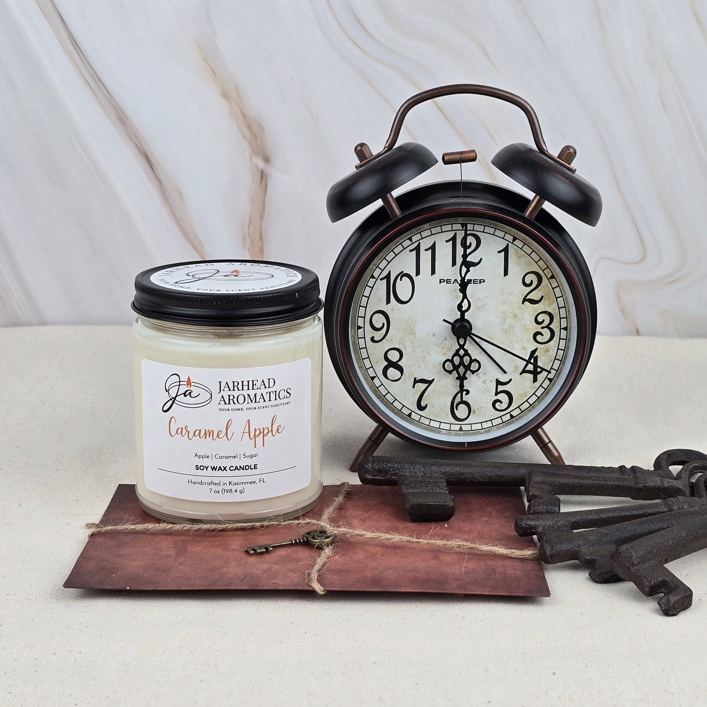 A black retro alarm clock reading 12:40 sits next to a Jarhead Aromatics Caramel Apple - Soy Wax Candle—evoking Granny Smith apples—plus a large vintage key and brown envelope on a light marble-patterned surface.