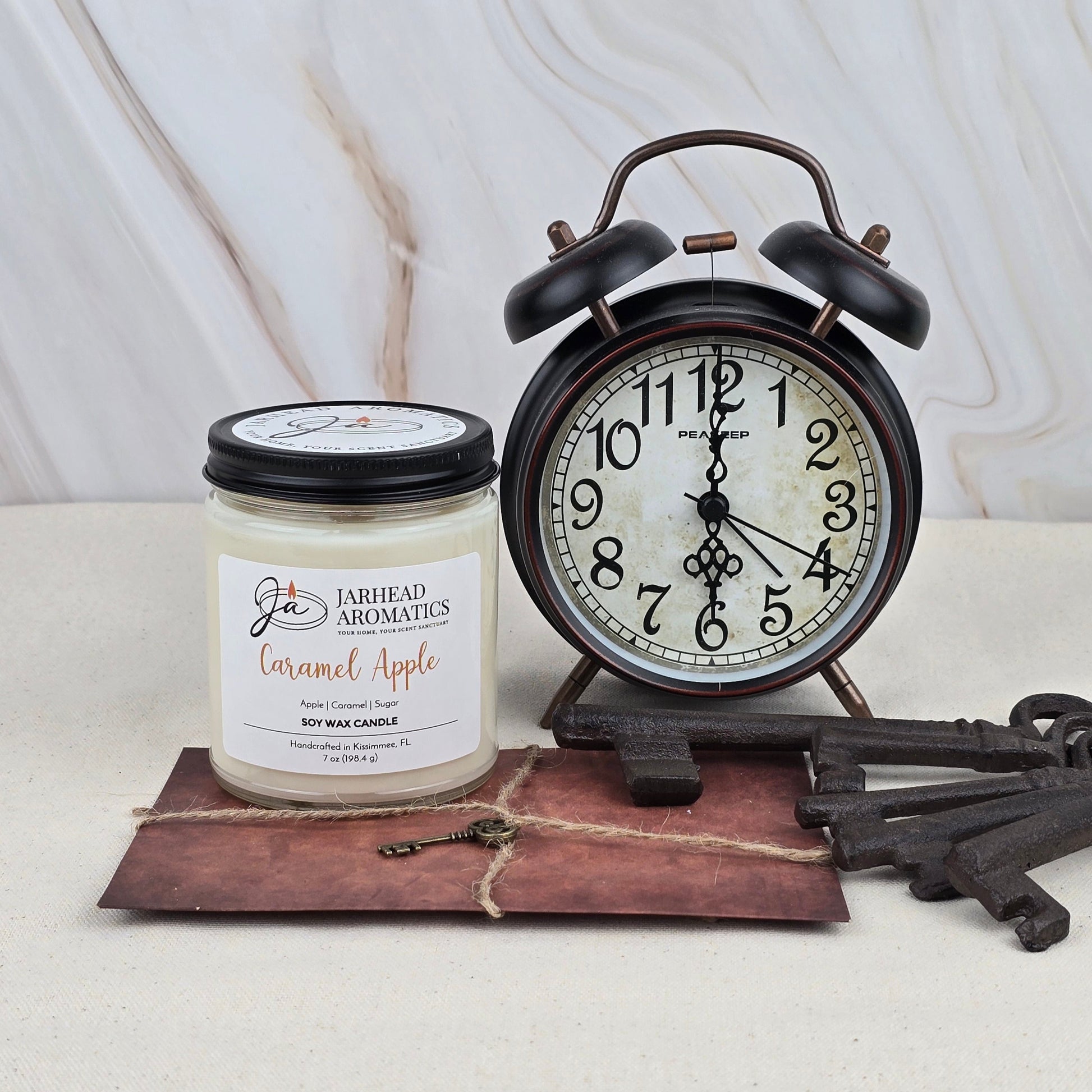 A black retro alarm clock reading 12:40 sits next to a Jarhead Aromatics Caramel Apple - Soy Wax Candle—evoking Granny Smith apples—plus a large vintage key and brown envelope on a light marble-patterned surface.