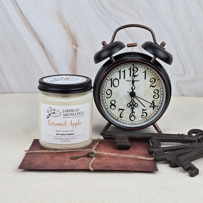 A black retro alarm clock reading 12:40 sits next to a Jarhead Aromatics Caramel Apple - Soy Wax Candle—evoking Granny Smith apples—plus a large vintage key and brown envelope on a light marble-patterned surface.