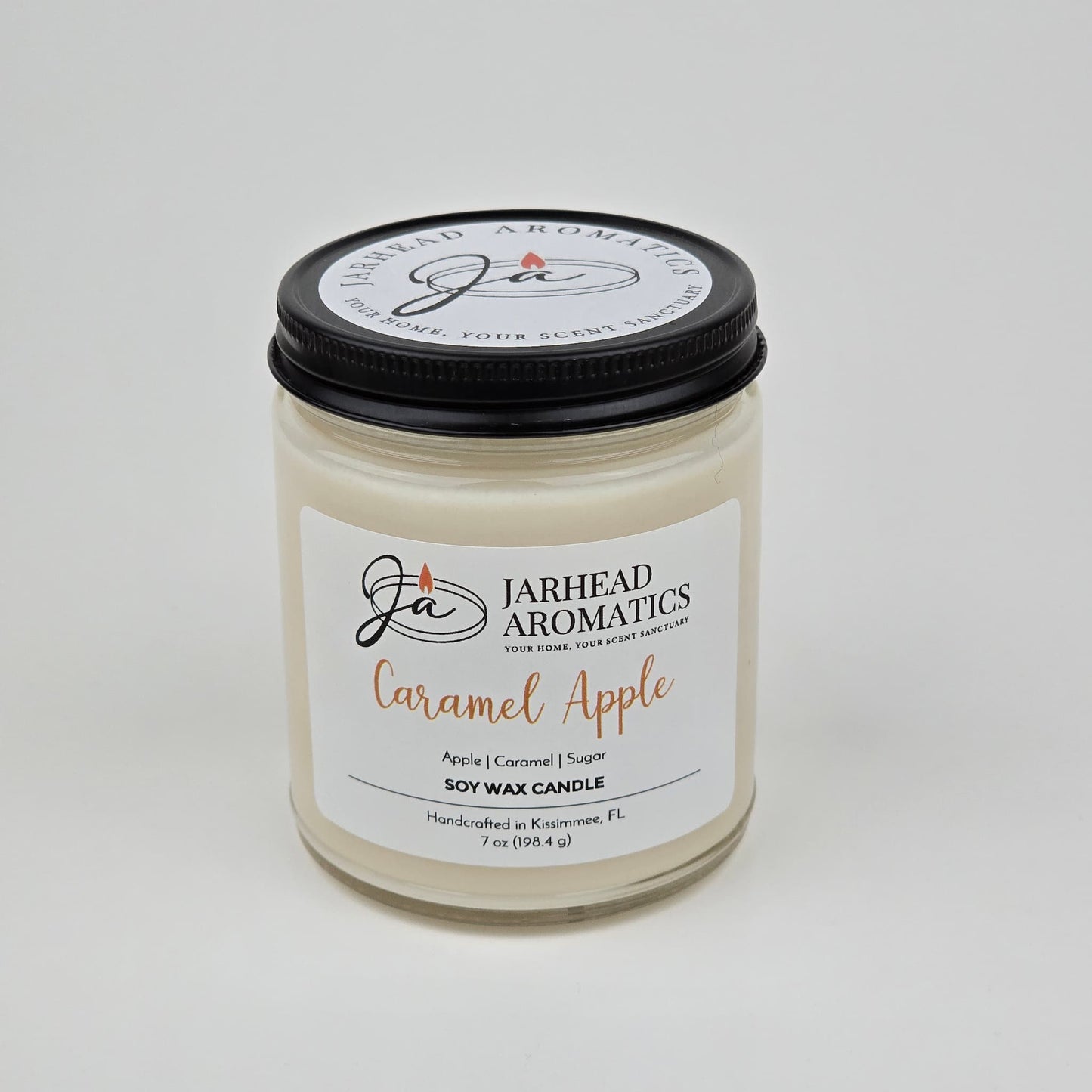 A Jarhead Aromatics Caramel Apple Soy Wax Candle, featuring a blend of apple cider, caramel, and sugar fragrances, is housed in a clear glass jar with a sleek black lid. The label emphasizes its enticing gourmand aroma and notes that it is handmade in Kissimmee, FL.