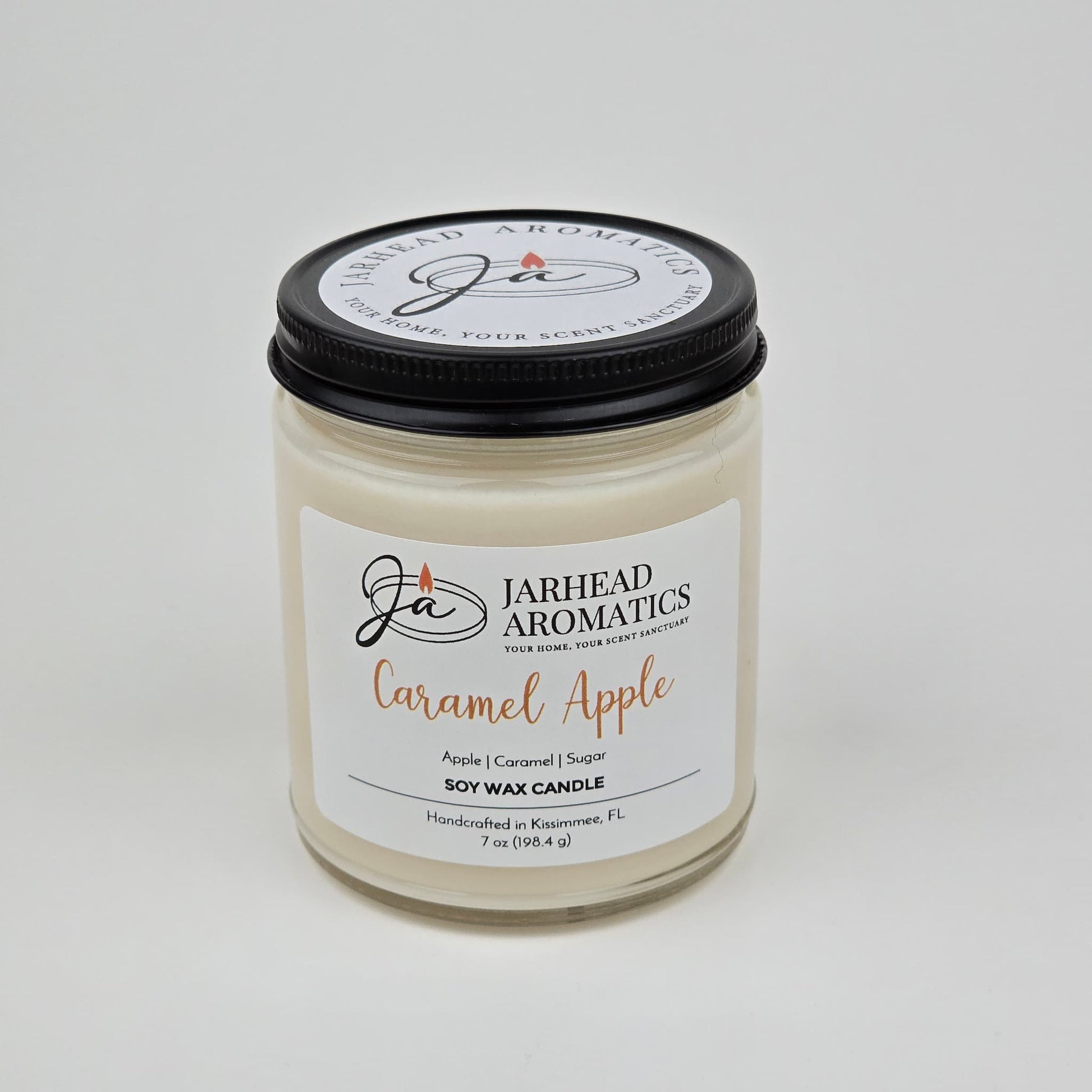 A Jarhead Aromatics Caramel Apple Soy Wax Candle, featuring a blend of apple cider, caramel, and sugar fragrances, is housed in a clear glass jar with a sleek black lid. The label emphasizes its enticing gourmand aroma and notes that it is handmade in Kissimmee, FL.