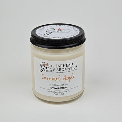 A Jarhead Aromatics Caramel Apple Soy Wax Candle, featuring a blend of apple cider, caramel, and sugar fragrances, is housed in a clear glass jar with a sleek black lid. The label emphasizes its enticing gourmand aroma and notes that it is handmade in Kissimmee, FL.