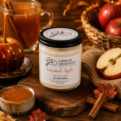 A Jarhead Aromatics Caramel Apple Soy Wax Candle | Apple Cider, Caramel & Sugar Candle rests on a wooden table surrounded by apples, a caramel apple, cinnamon sticks, caramel sauce, and a mug of apple cider, creating a warm and cozy autumn ambiance.