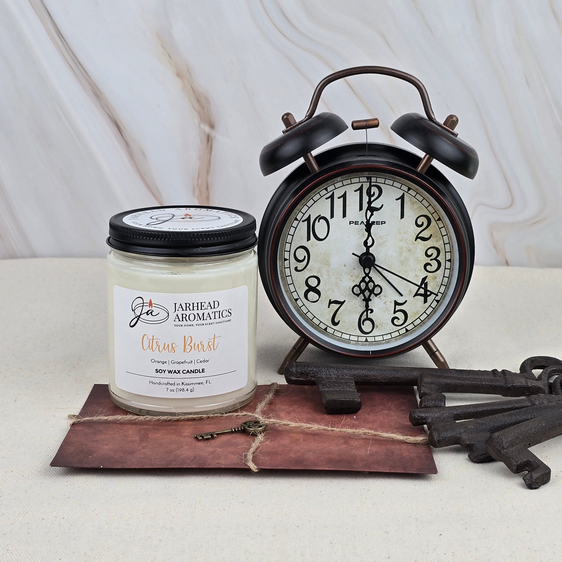 A Citrus Burst - Soy Wax Candle from Jarhead Aromatics, made with essential oils, is placed beside a vintage black alarm clock, antique keys, and a brown tied envelope on a light surface with a marble background.