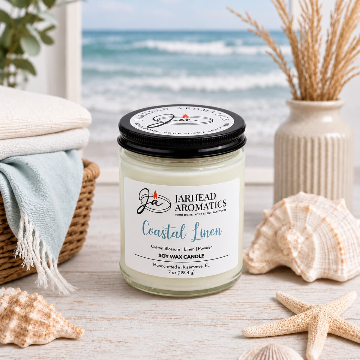 A Jarhead Aromatics Coastal Linen Soy Wax Candle in a glass jar sits on a wooden surface surrounded by seashells and a basket of towels, with a beach scene in the background.