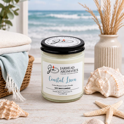 A Jarhead Aromatics Coastal Linen Soy Wax Candle in a glass jar sits on a wooden surface surrounded by seashells and a basket of towels, with a beach scene in the background.