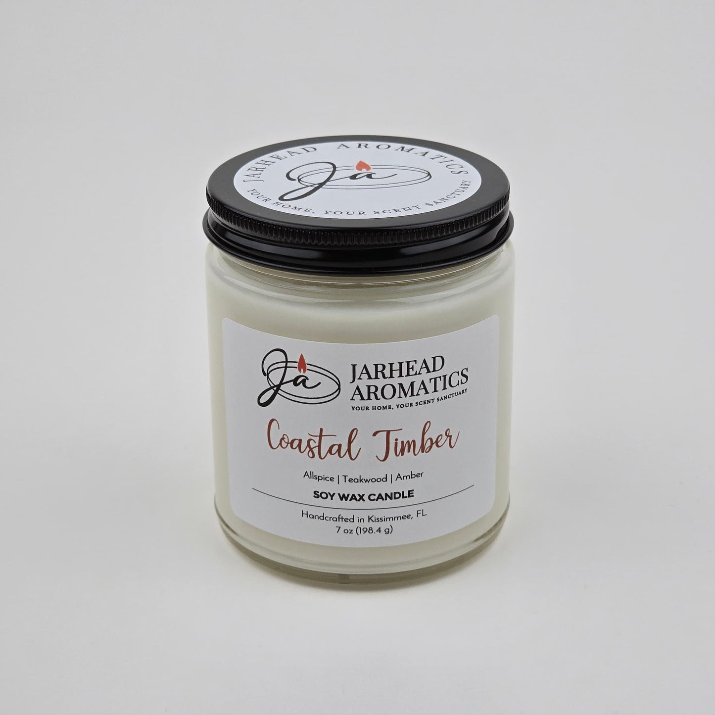 A white premium soy wax candle in a glass jar with a black lid labeled Jarhead Aromatics Coastal Timber Soy Wax Candle, highlighting masculine notes of teakwood, leather, and amber. The label indicates it is handcrafted in Kissimmee, FL.