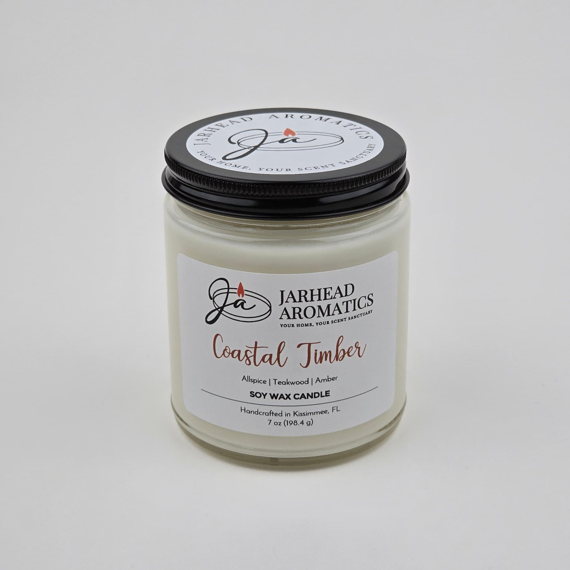 A white premium soy wax candle in a glass jar with a black lid labeled Jarhead Aromatics Coastal Timber Soy Wax Candle, highlighting masculine notes of teakwood, leather, and amber. The label indicates it is handcrafted in Kissimmee, FL.
