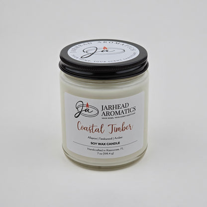 A white premium soy wax candle in a glass jar with a black lid labeled Jarhead Aromatics Coastal Timber Soy Wax Candle, highlighting masculine notes of teakwood, leather, and amber. The label indicates it is handcrafted in Kissimmee, FL.