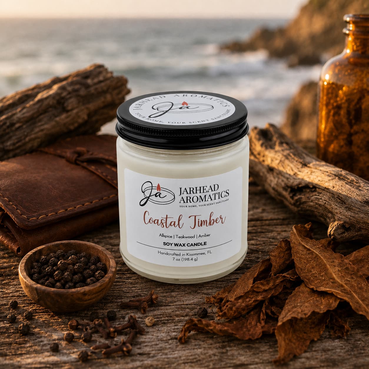A Jarhead Aromatics Coastal Timber Soy Wax Candle | Teakwood, Leather & Amber Candle sits on a wooden surface alongside peppercorns, tobacco leaves, driftwood, and a brown notebook, its masculine blend of teakwood, leather, and amber evoking the rugged ocean and rocky shore in the background.