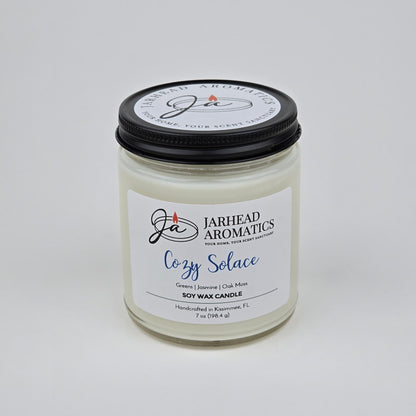 The Cozy Solace Soy Wax Candle by Jarhead Aromatics features fresh greens, jasmine, oak moss, and musk—an ideal fall scent—and comes hand-poured in a clear glass jar with a black lid.