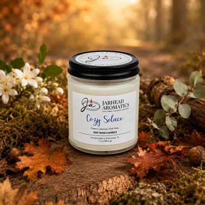 A Jarhead Aromatics Cozy Solace Soy Wax Candle—with notes of fresh greens, jasmine, oak moss, and musk—rests on a log among autumn leaves and flowers in a sunlit forest setting, infusing the air with its welcoming fall fragrance.