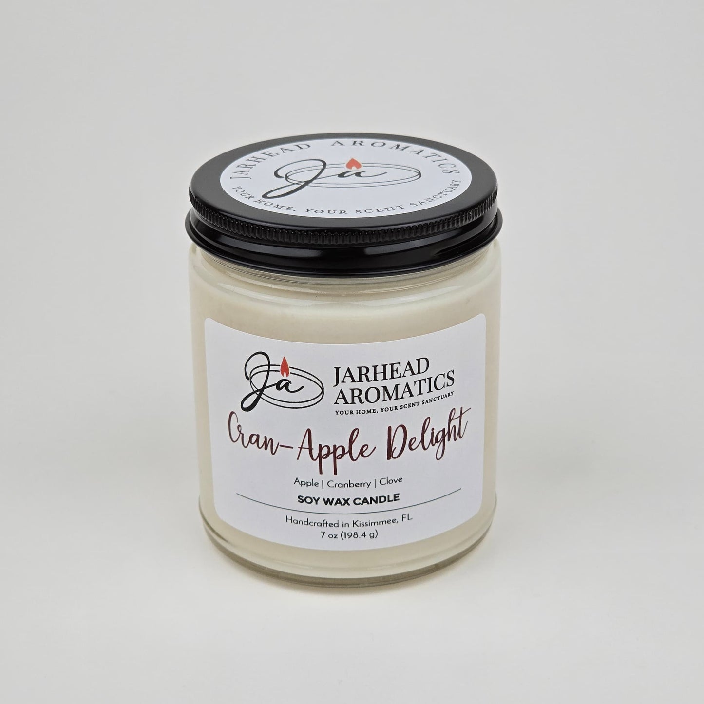 A Jarhead Aromatics Cran-Apple Delight Soy Wax Candle, featuring the scent of cranberry, apple, and cinnamon for a long-lasting fragrance, is displayed in a clear jar with a black lid against a plain white background.