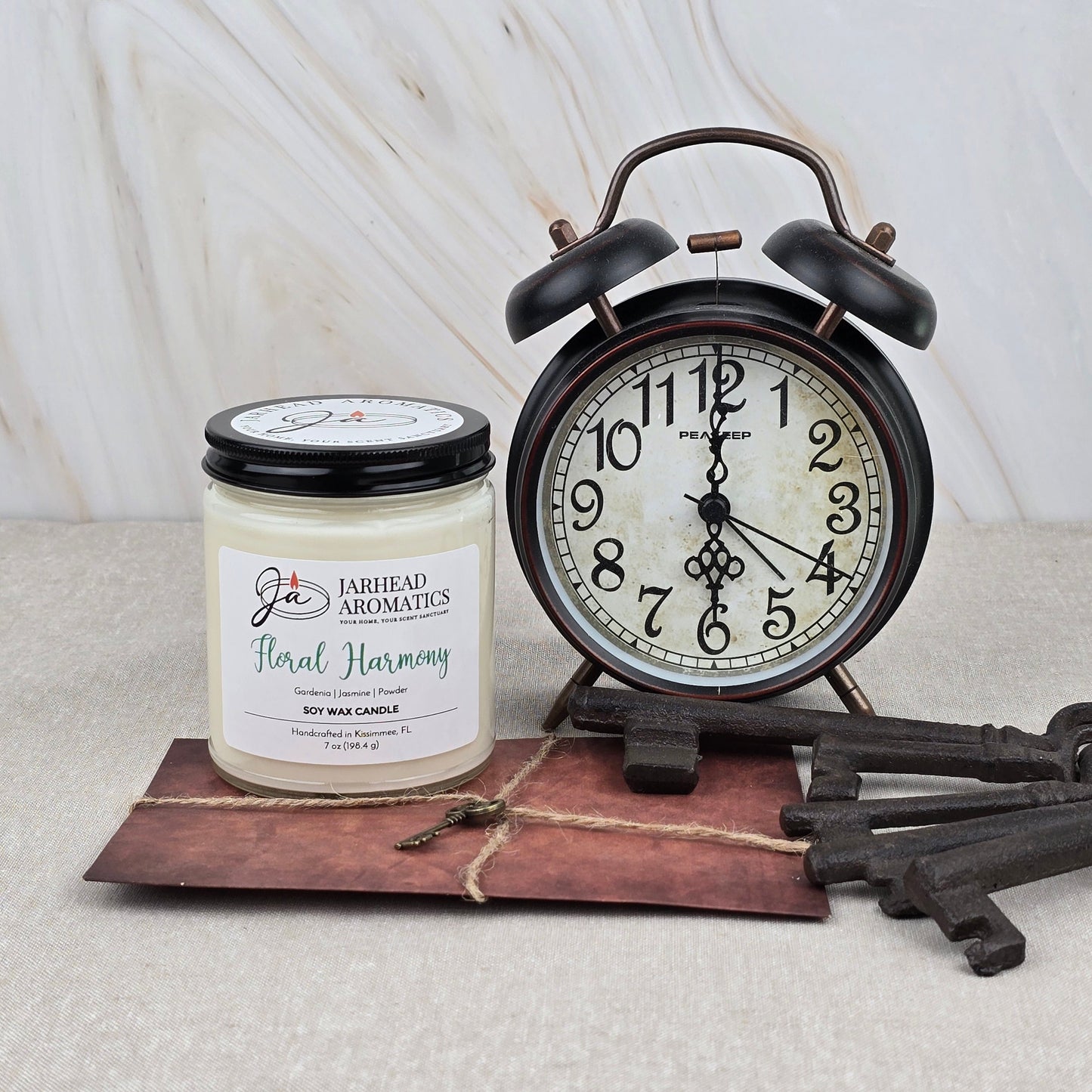 A Floral Harmony Soy Wax Candle by Jarhead Aromatics, scented with gardenia, jasmine & tuberose, rests on a brown envelope with three vintage keys beside a classic black twin-bell clock showing 9:07 on a light marble background.