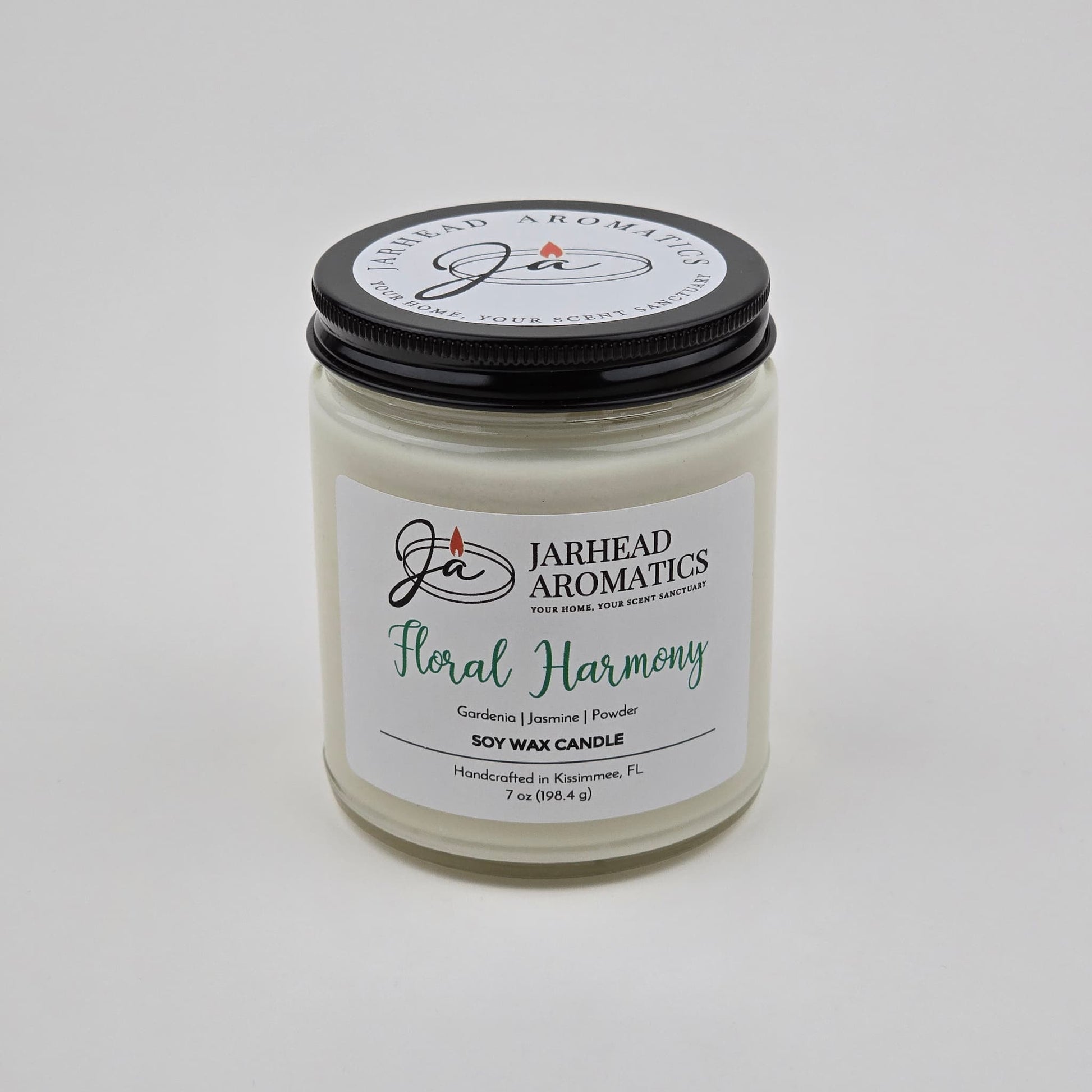 Experience the Floral Harmony Soy Wax Candle by Jarhead Aromatics—a hand-poured candle crafted in Kissimmee, FL. Presented in a clear jar with a black lid featuring the brand logo and text, this candle blends gardenia, jasmine, and tuberose for a long-lasting fragrance that infuses your space with delicate white floral notes.