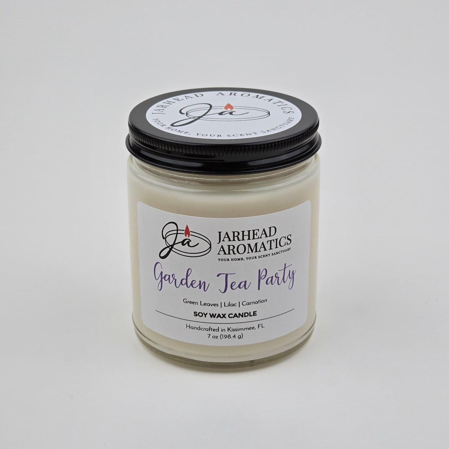A white soy wax candle in a clear glass jar with a black lid. The label reads Jarhead Aromatics Garden Tea Party Soy Wax Candle | Rose, Jasmine & Green Floral Candle. This handcrafted floral scented candle, featuring notes of rose, jasmine, and green florals, is made by Jarhead Aromatics in Kissimmee, FL.