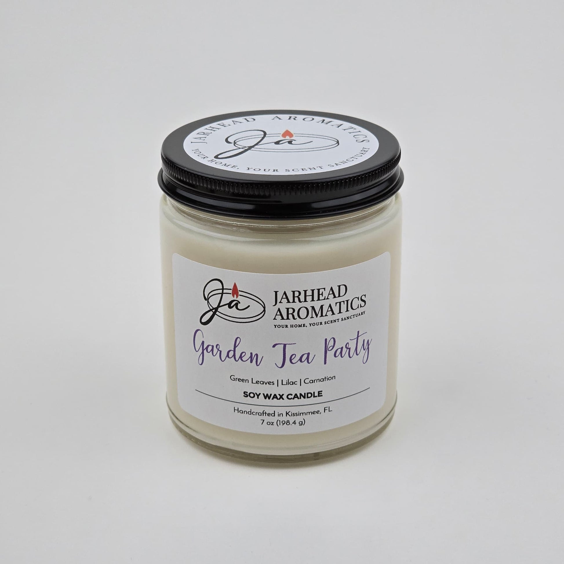 A white soy wax candle in a clear glass jar with a black lid. The label reads Jarhead Aromatics Garden Tea Party Soy Wax Candle | Rose, Jasmine & Green Floral Candle. This handcrafted floral scented candle, featuring notes of rose, jasmine, and green florals, is made by Jarhead Aromatics in Kissimmee, FL.