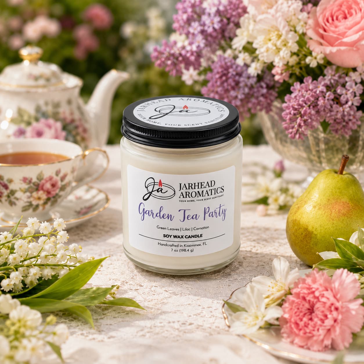A Jarhead Aromatics Garden Tea Party Soy Wax Candle, featuring notes of rose, jasmine, and green florals, sits on a table with flowers, a teacup and saucer, a pear, and a floral teapot—evoking the ambiance of a spring garden tea party.