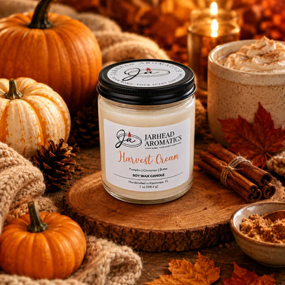 A Jarhead Aromatics Harvest Cream Soy Wax Candle—featuring the inviting scents of pumpkin spice, brown sugar, and vanilla—rests on a wood slab surrounded by pumpkins, pinecones, a cozy sweater, cinnamon sticks, a latte, and autumn leaves in warm golden light, filling your home with the perfect fall fragrance.