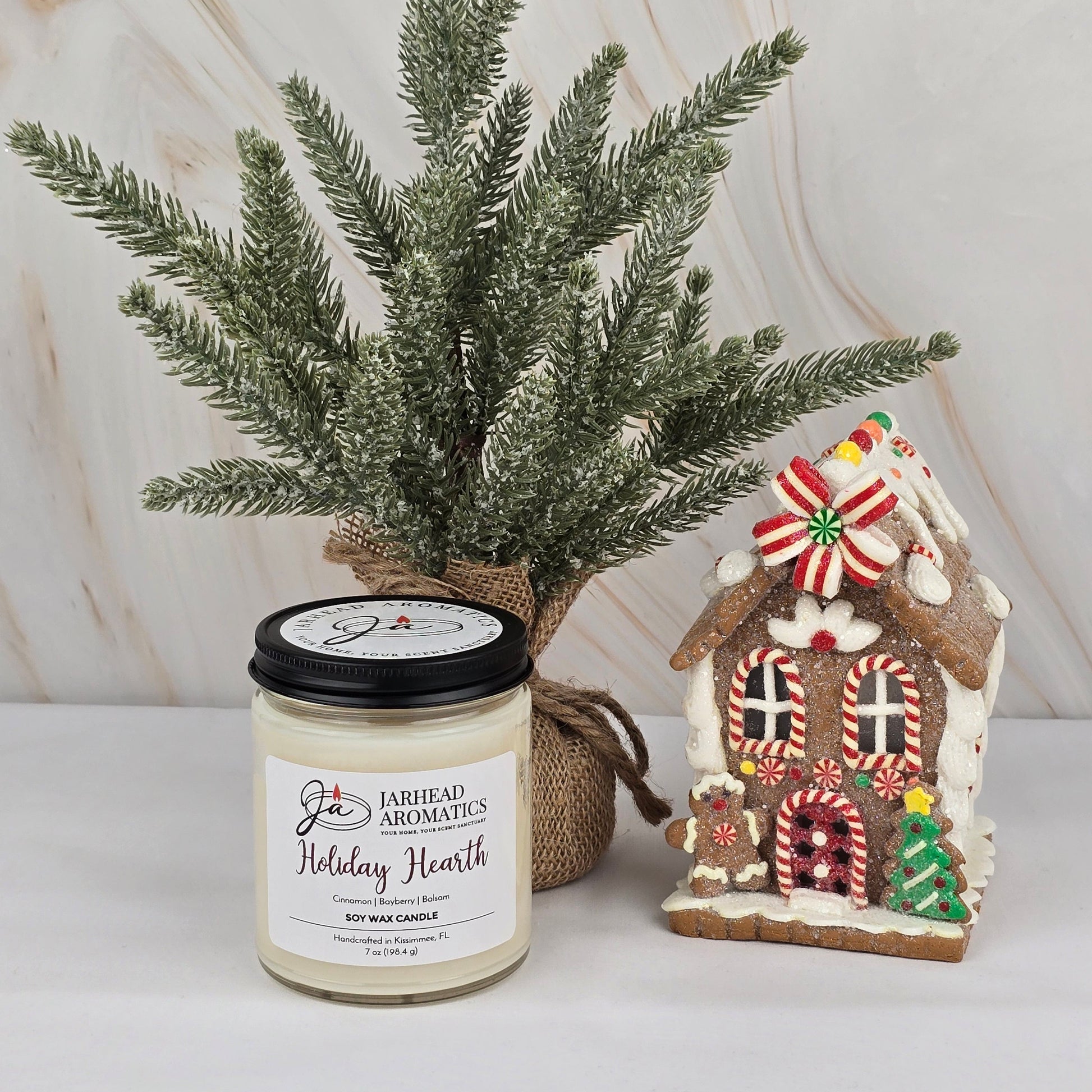 A small pine tree in burlap, Jarhead Aromatics’ Holiday Hearth Soy Wax Candle (Winter Collection), and a decorated gingerbread house ornament create a cozy holiday vibe on a light surface with a marble backdrop.