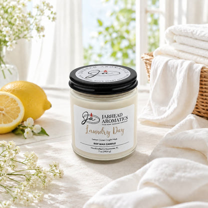 A Laundry Day Soy Wax Candle | Fresh Linen, Lemon & Light Musk Candle from Jarhead Aromatics rests on a white surface surrounded by fresh lemons, folded towels, a wicker basket, and delicate white flowers in a bright, sunlit room. This clean linen candle fills your space with a long-lasting fragrance that embodies freshness and comfort.