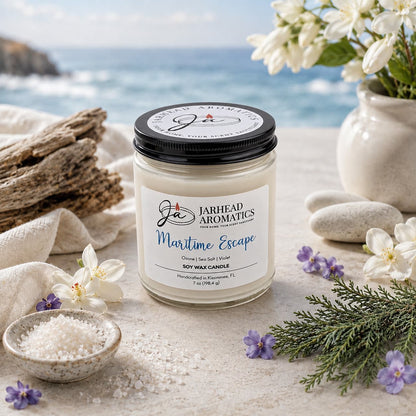 A jar candle from Jarhead Aromatics, labeled Maritime Escape Soy Wax Candle | Sea Salt, Ozone & Cedar Candle, rests on a stone surface surrounded by flowers, sea salt, driftwood, and a view of the ocean. Infused with notes of sea salt, ozone, and cedar, this coastal candle offers an ocean-inspired fragrance and is made from premium soy wax for a clean and relaxing burn.