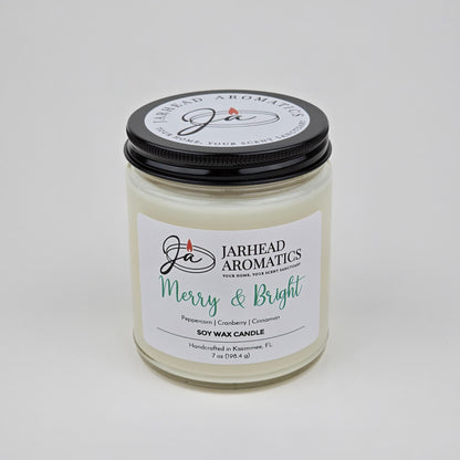 A white Merry & Bright Soy Wax Candle by Jarhead Aromatics with a black lid is displayed against a plain white background. The label reads "Merry & Bright" and features notes of citrus cranberry, warm spice, and fir—capturing the essence of a festive holiday fragrance.