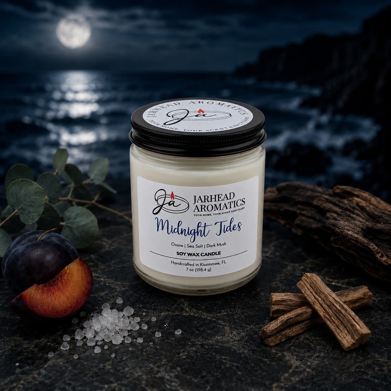 A Midnight Tides Soy Wax Candle by Jarhead Aromatics rests on a dark surface surrounded by eucalyptus leaves, plum, sea salt, and wood, as a moonlit ocean and rocky shore unfold in the background. Hand-poured and scented with sea salt, dark musk, and sandalwood, this candle captures the essence of coastal evenings with its enchanting aroma.