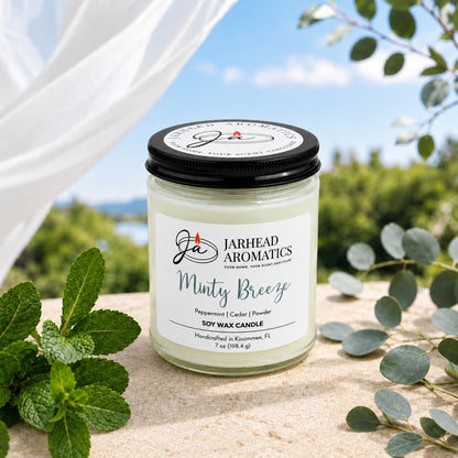 A Jarhead Aromatics Minty Breeze Soy Wax Candle in a glass jar sits on a stone surface surrounded by mint leaves and eucalyptus branches.
