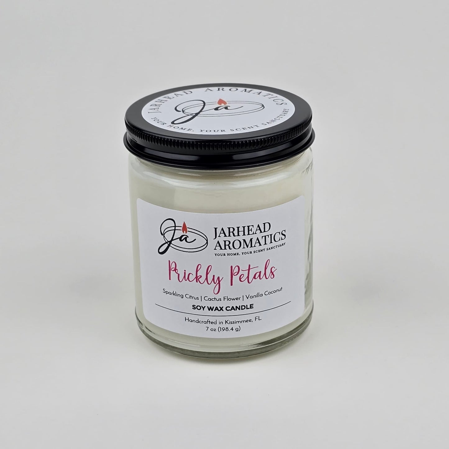 The Prickly Petals Soy Wax Candle by Jarhead Aromatics features cactus flower and citrus scents with notes of vanilla coconut. Handcrafted in Kissimmee, FL, this desert-inspired candle comes in a glass jar with a black lid and white wax.