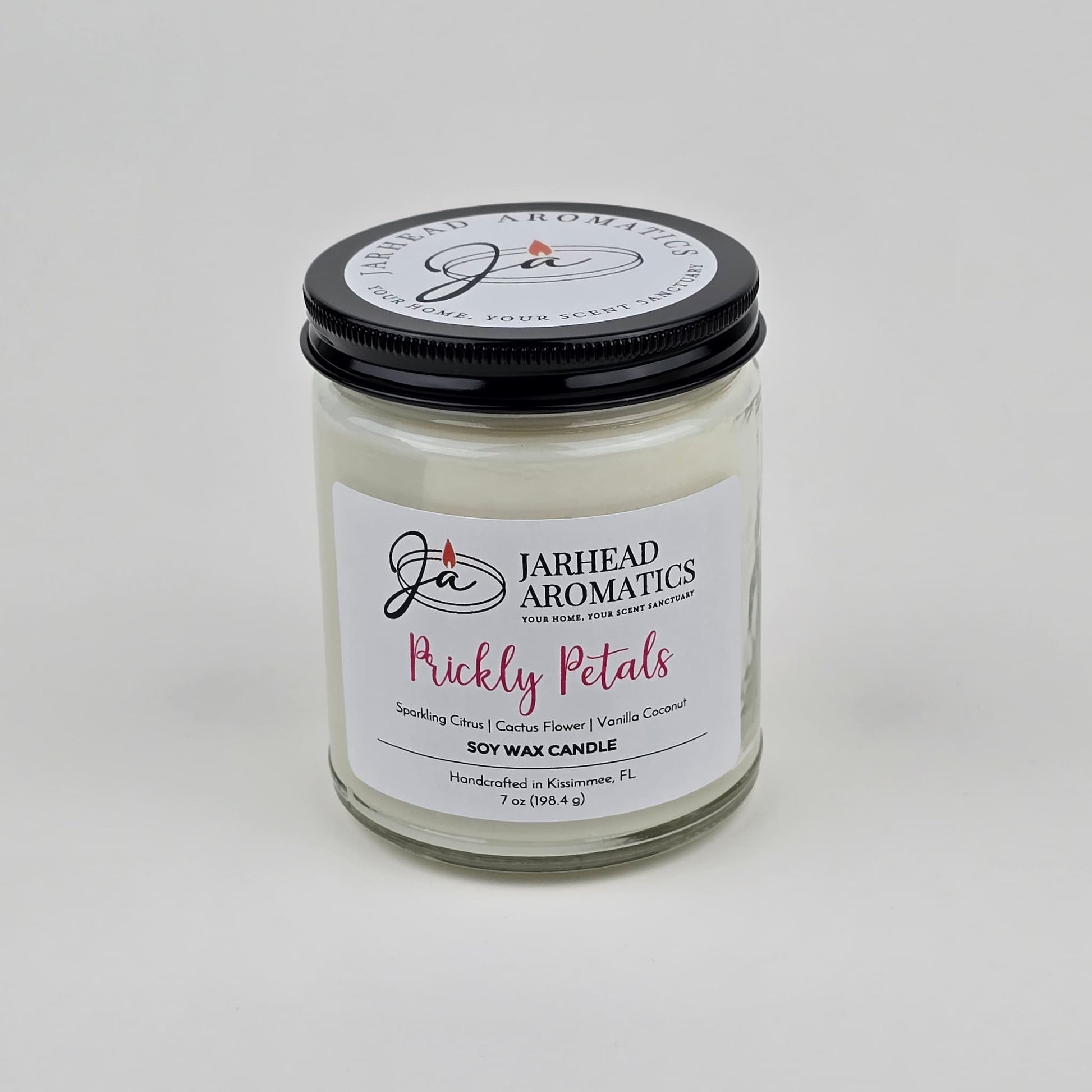 The Prickly Petals Soy Wax Candle by Jarhead Aromatics features cactus flower and citrus scents with notes of vanilla coconut. Handcrafted in Kissimmee, FL, this desert-inspired candle comes in a glass jar with a black lid and white wax.
