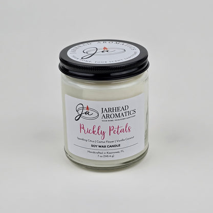 The Prickly Petals Soy Wax Candle by Jarhead Aromatics features cactus flower and citrus scents with notes of vanilla coconut. Handcrafted in Kissimmee, FL, this desert-inspired candle comes in a glass jar with a black lid and white wax.