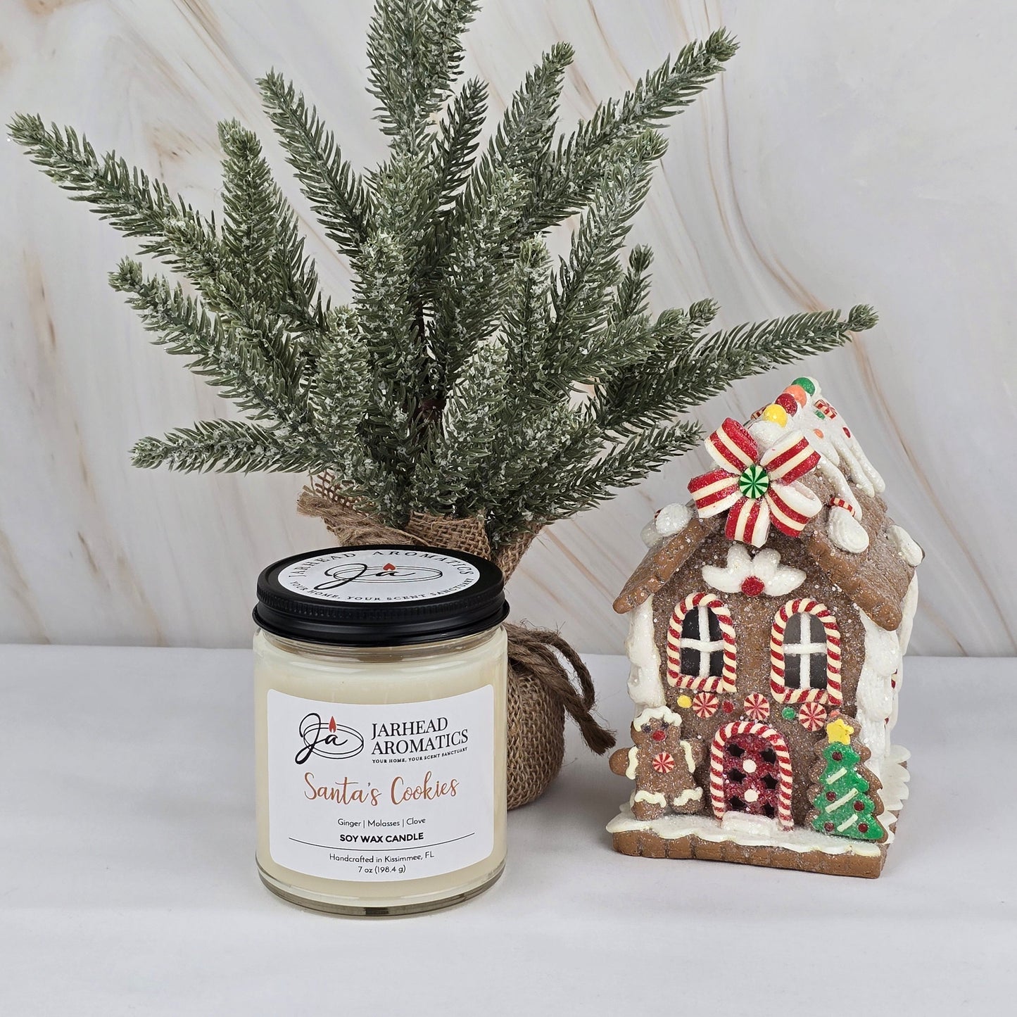 A small frosted pine tree, a Jarhead Aromatics Santa’s Cookies - Soy Wax Candle from the Winter Collection, and a decorated gingerbread house are displayed on a white surface against a light background.