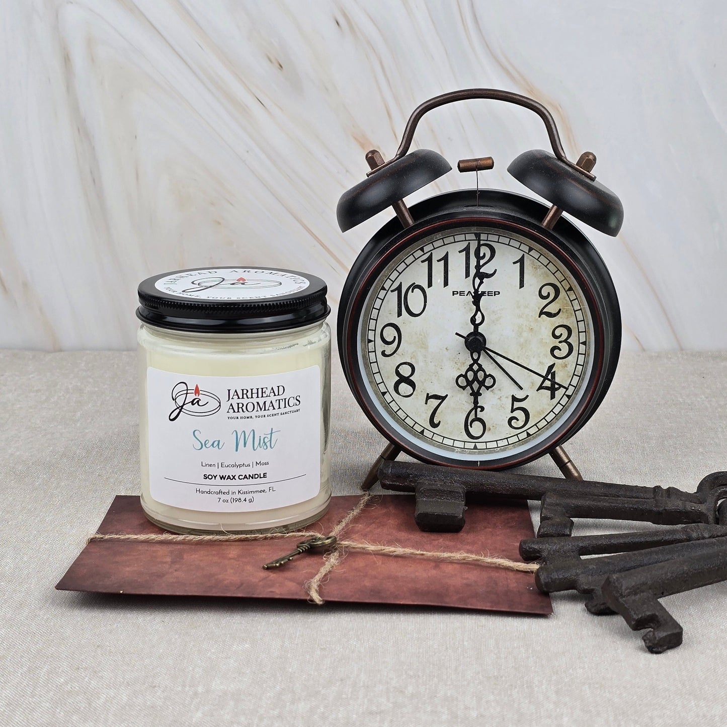 A vintage black alarm clock, a Jarhead Aromatics Sea Mist Soy Wax Candle in Clean Coastal Linen & Sea Salt scent, a brown envelope tied with twine, and three large metal keys are arranged on a light marble surface.