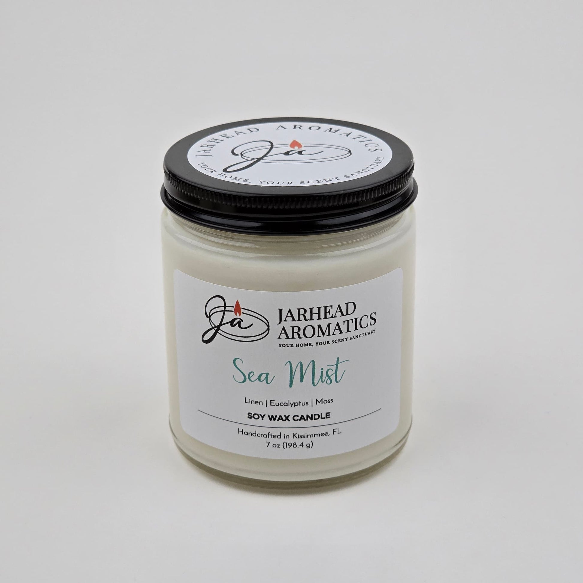 A white glass jar candle labeled Jarhead Aromatics Sea Mist Soy Wax Candle | Clean Coastal Linen & Sea Salt Candle, featuring notes of linen, eucalyptus, and moss. Handcrafted in Kissimmee, FL, this coastal candle offers an aquatic floral fragrance and comes topped with a black lid.