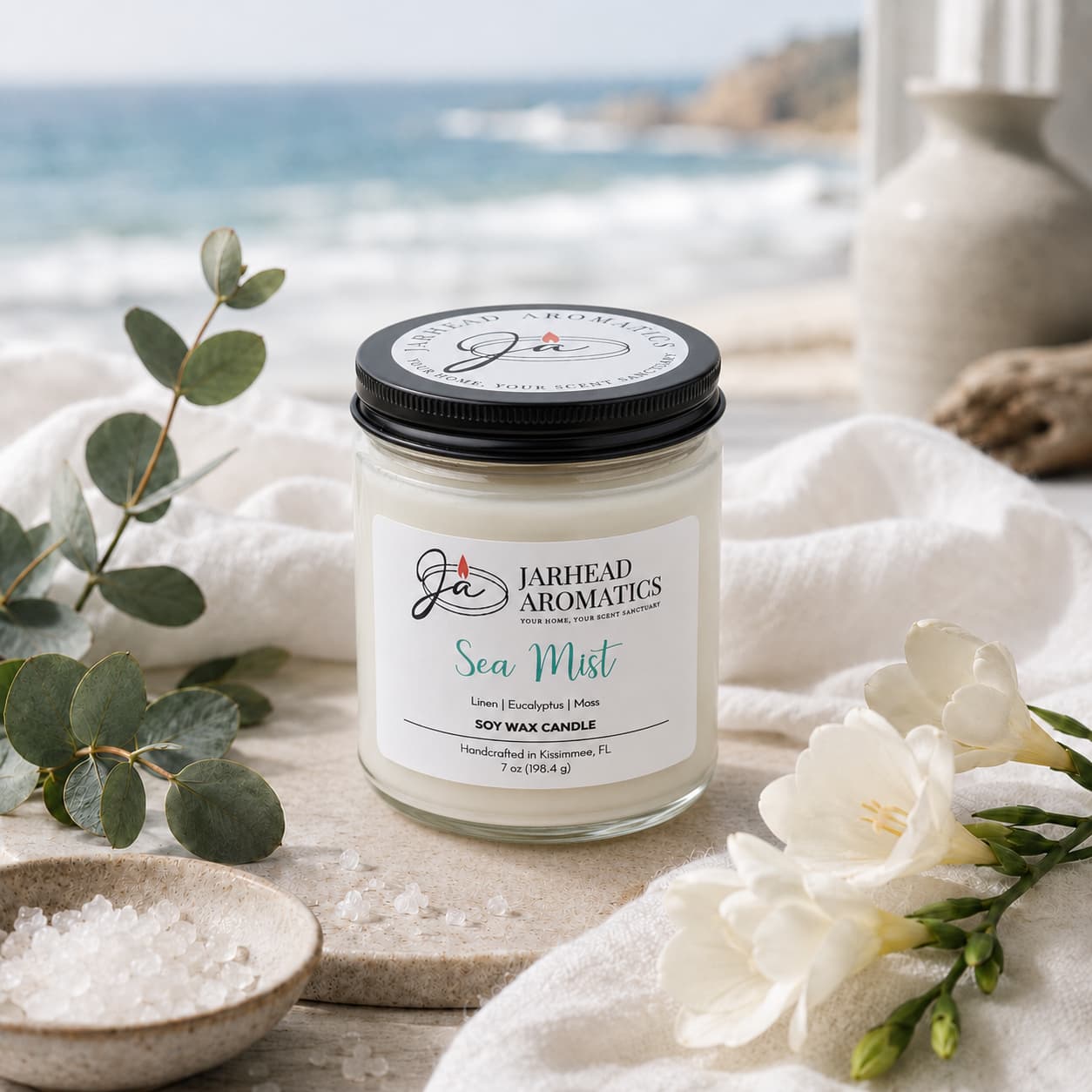 A Jarhead Aromatics Sea Mist Soy Wax Candle | Clean Coastal Linen & Sea Salt Candle rests on a white cloth beside sea salt, eucalyptus, and white flowers, infusing the air with its rejuvenating aquatic floral aroma against the tranquil backdrop of a serene coastal view and a blurred beach.