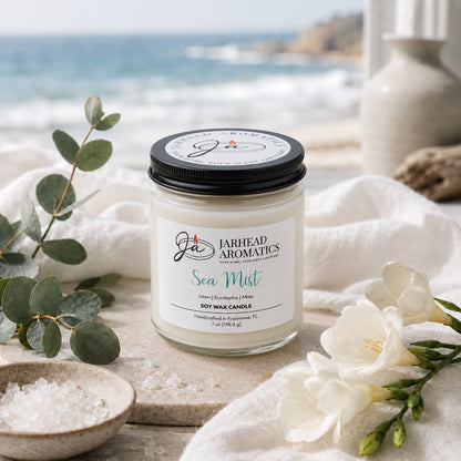 A Jarhead Aromatics Sea Mist Soy Wax Candle | Clean Coastal Linen & Sea Salt Candle rests on a white cloth beside sea salt, eucalyptus, and white flowers, infusing the air with its rejuvenating aquatic floral aroma against the tranquil backdrop of a serene coastal view and a blurred beach.
