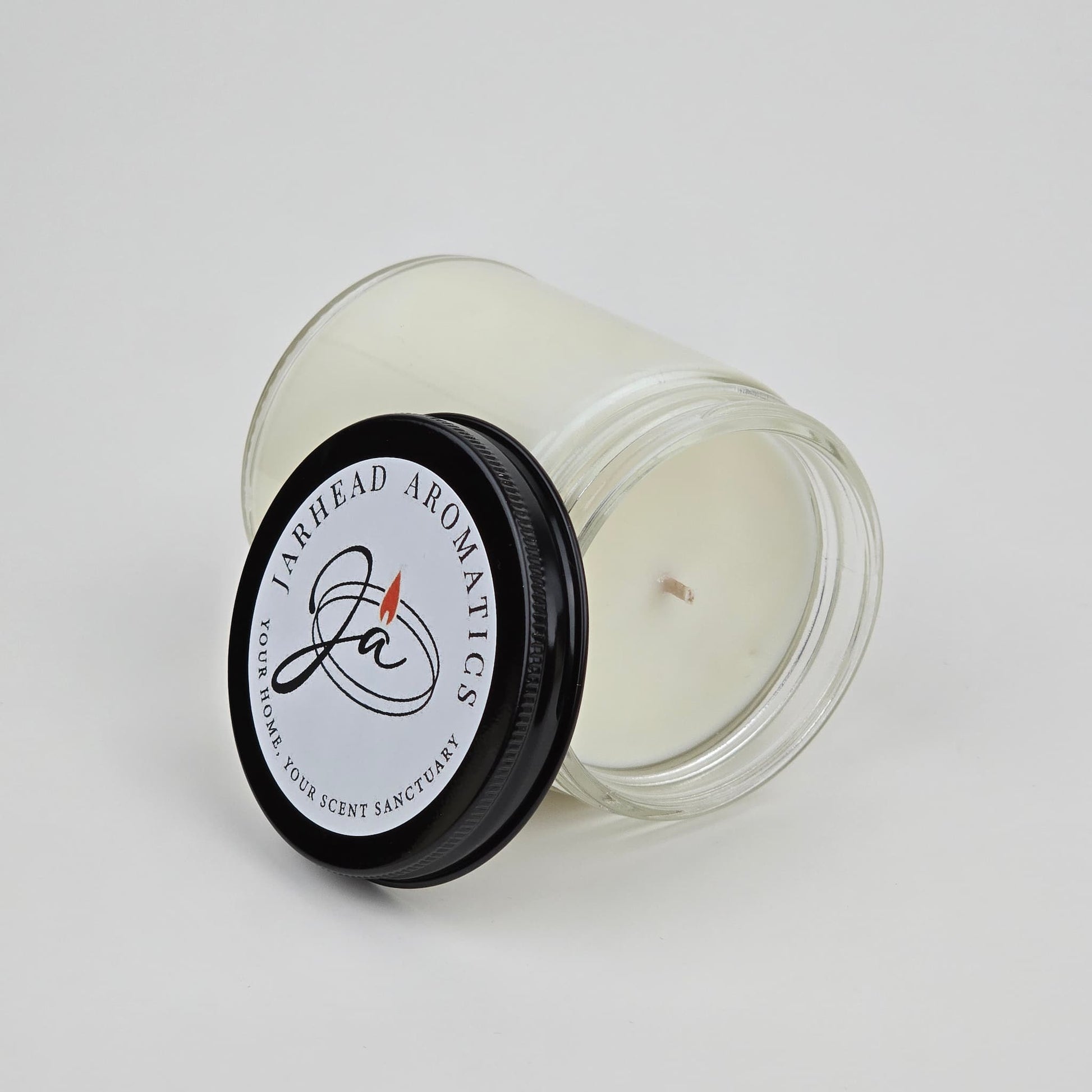 A white candle, The Lumberjack Soy Wax Candle by Jarhead Aromatics, rests in a clear glass jar with its black logo lid nearby, suggesting a masculine cedar, oak & mahogany scent inspired by a woodland cabin sanctuary.